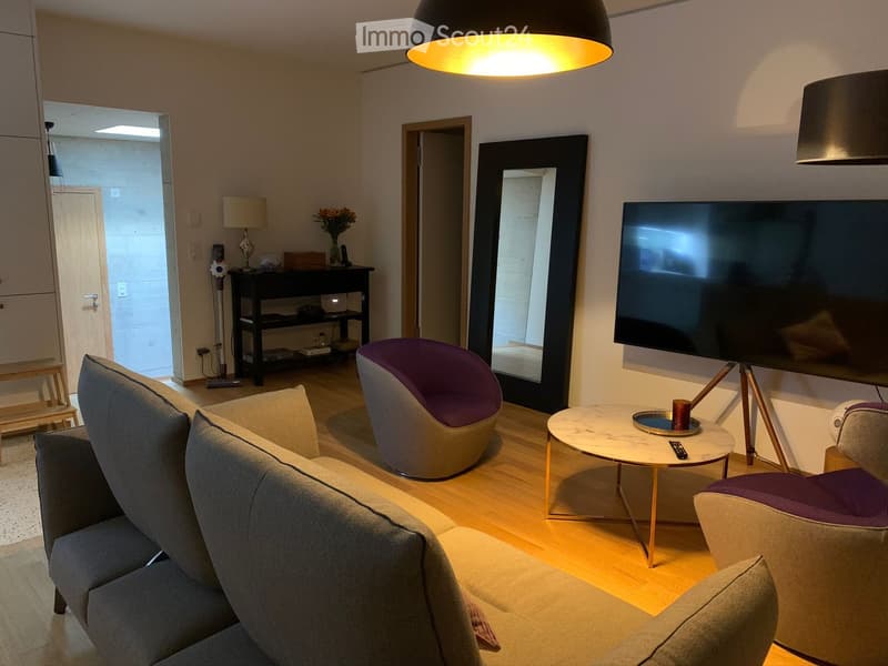 Modern living room with a grey sofa, coffee table, two chairs, a large mirror, and a television mounted on the wall. The room features wooden flooring and warm lighting from a hanging lamp.