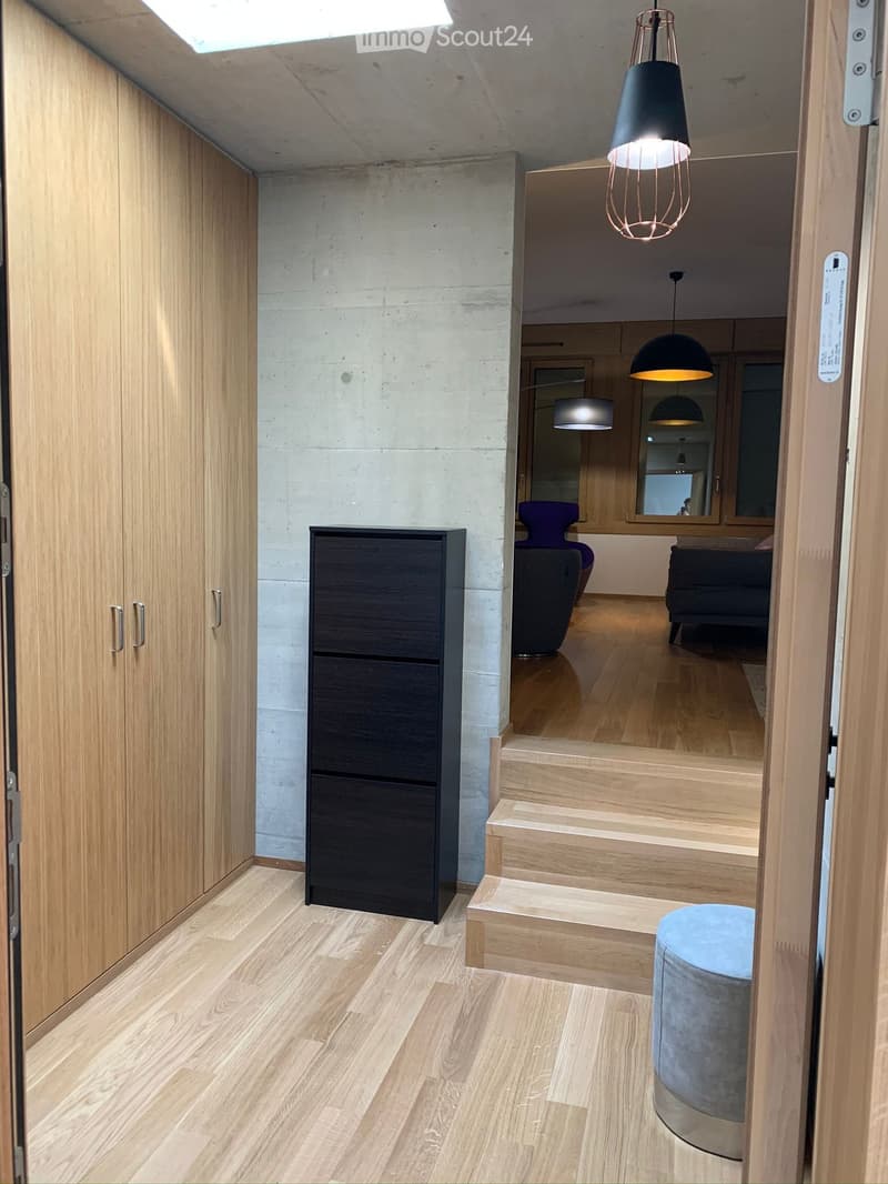 Modern hallway with wooden flooring, built-in wooden cabinets, a small black storage unit, and a staircase leading to another level. A round blue ottoman is placed near the stairs.