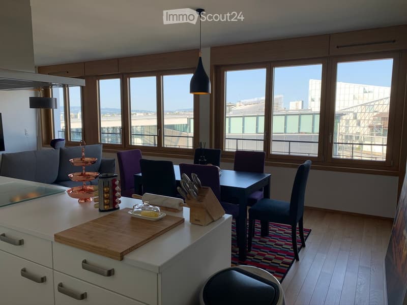 Modern open-plan living and dining area with large windows offering a city view. Features a grey sofa, dining table with chairs, kitchen island with cutting board and knife block, and pendant lighting.