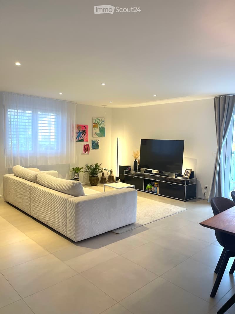 Modern living room with a white sectional sofa, coffee table, large flat-screen TV, and decorative vases. The space features large windows with sheer curtains and a tiled floor.