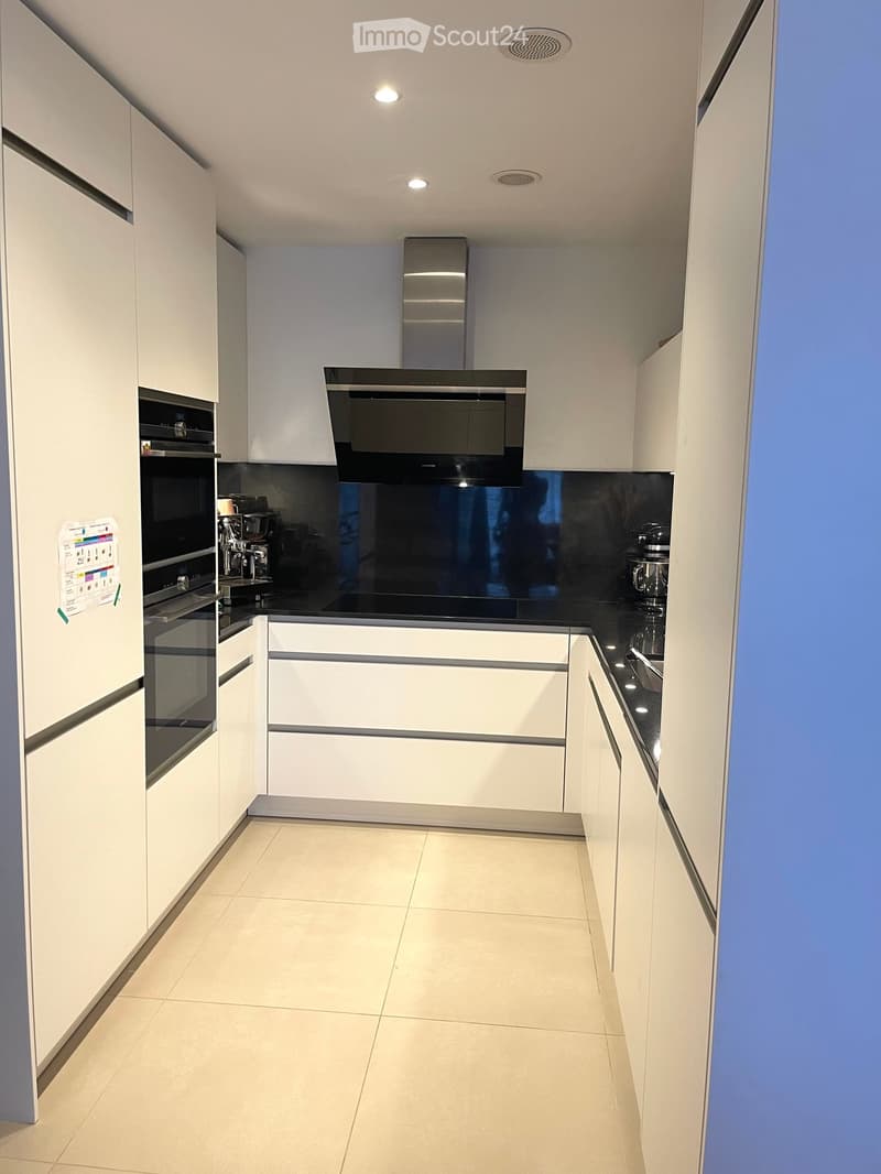 Modern kitchen with white cabinets, black countertops, and stainless steel appliances including an oven, coffee machine, and range hood.