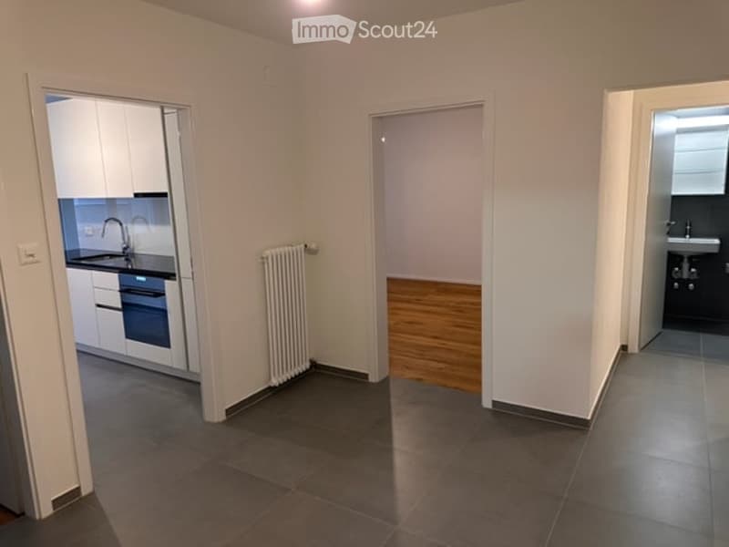 Empty room with wooden flooring, white walls, and a radiator. Adjacent rooms include a kitchen and a bathroom.