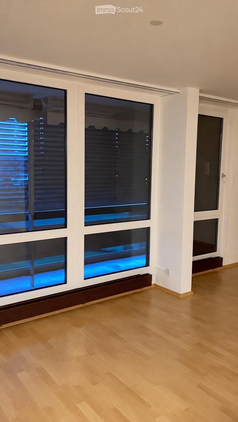 Empty room with light wooden flooring, white walls, large windows with blinds, and a small radiator under the windows.