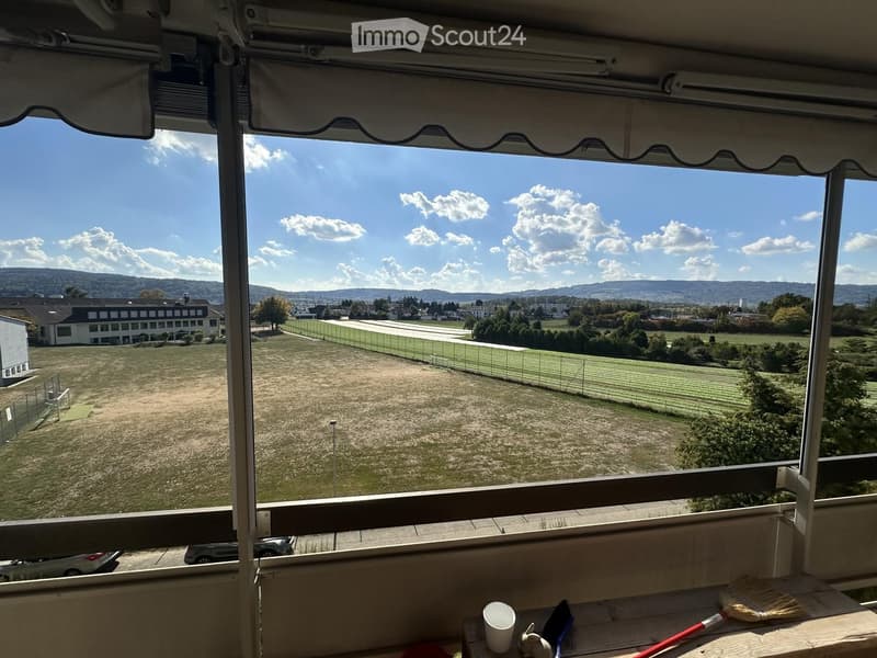 Balcony with a retractable awning, overlooking a grassy field, buildings, and distant hills under a partly cloudy sky.