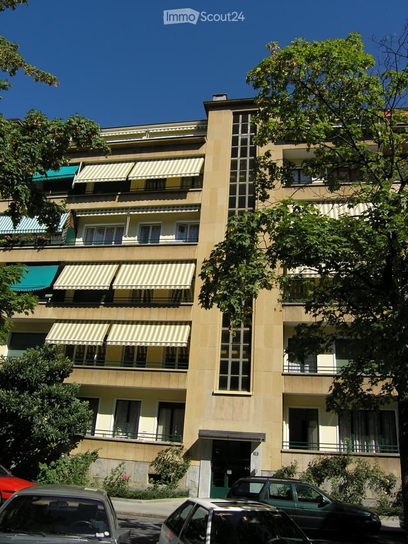 Multi-story residential building with beige facade, balconies, striped awnings, and surrounding greenery.