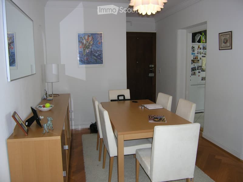 Dining room with a wooden dining table, six white chairs, a sideboard with a lamp, a mirror, and a refrigerator in the background.