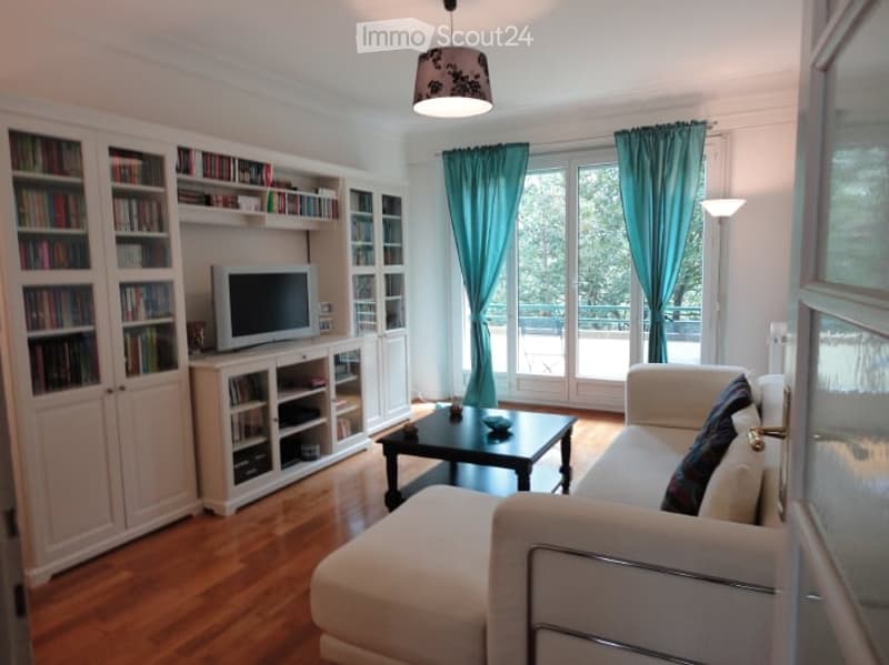 Bright living room with large windows and a view, featuring a white sectional sofa, a black coffee table, a television on a white cabinet, built-in bookshelves, and a floor lamp.