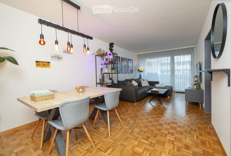 Modern living and dining area with wooden flooring, a grey sofa, a dining table with chairs, and decorative shelves.