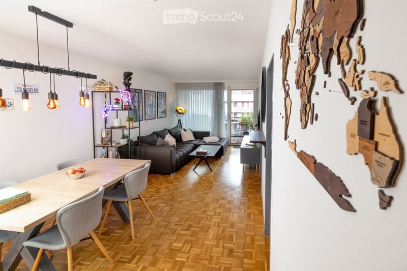Modern living and dining area with wooden flooring, a grey sectional sofa, a coffee table, a dining table with chairs, and a decorative wall map.