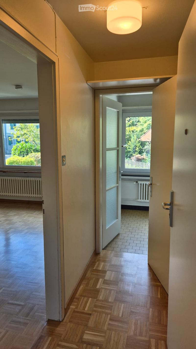 A narrow hallway with wooden flooring, light-colored walls, and a ceiling light fixture. Open doors lead to adjacent rooms with visible windows and radiators.