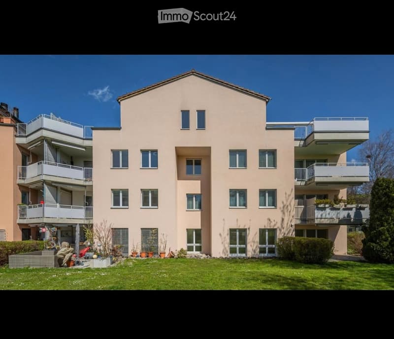 A modern, multi-story residential building with light beige walls, multiple windows, and balconies. The building is surrounded by a well-maintained lawn and small garden area with potted plants.