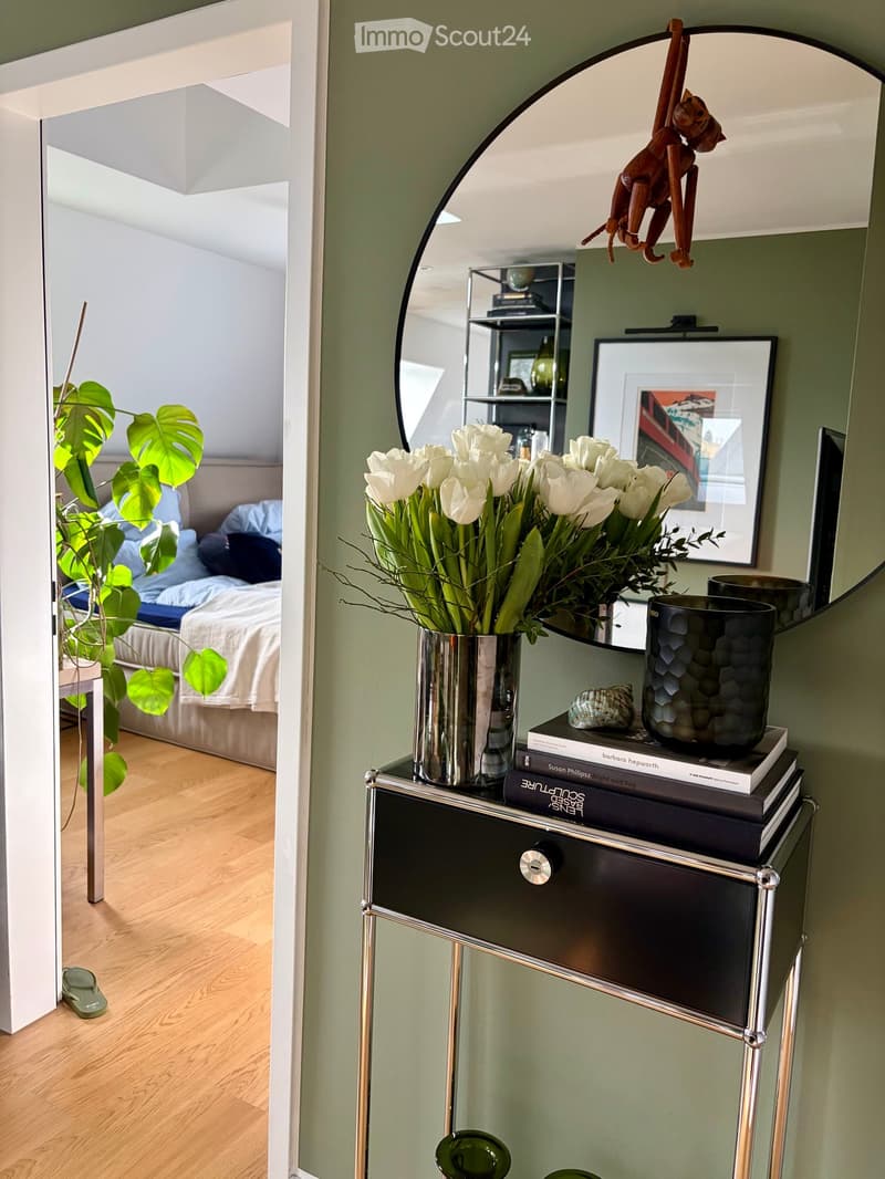 Modern bedroom with a bed, nightstand, mirror, vase with white flowers, books, and a plant in the corner.
