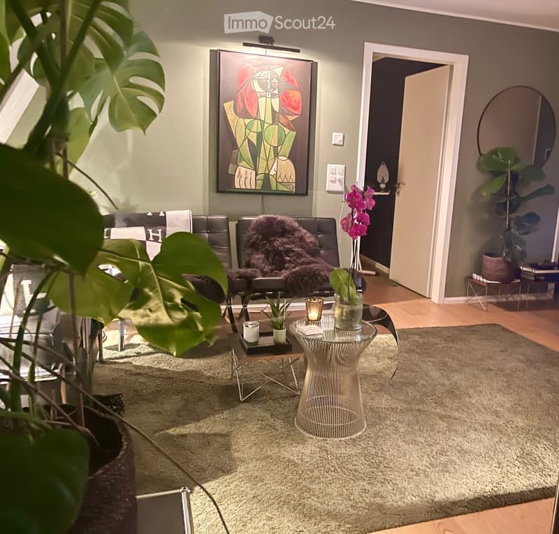 Modern living room with a large abstract painting, a round glass coffee table, a wicker side table, a plush chair with a fur throw, and a large potted plant in the foreground.