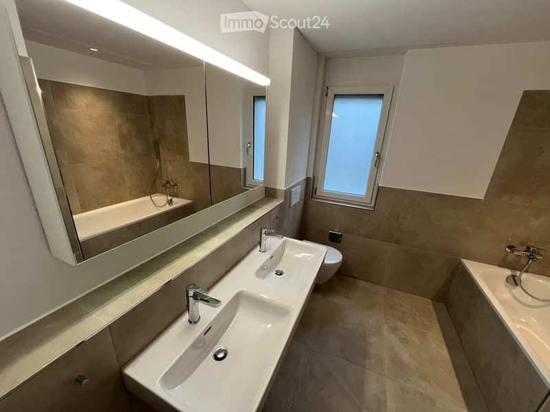 Modern bathroom with double sink, large mirror, bathtub, and toilet. Features two windows and sleek fixtures.