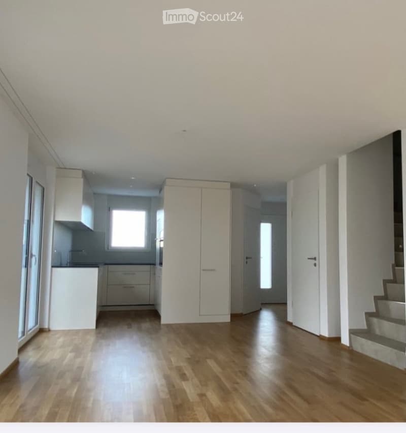 Empty room with wooden flooring, white walls, and a small kitchen area visible in the background. No furniture or equipment present.