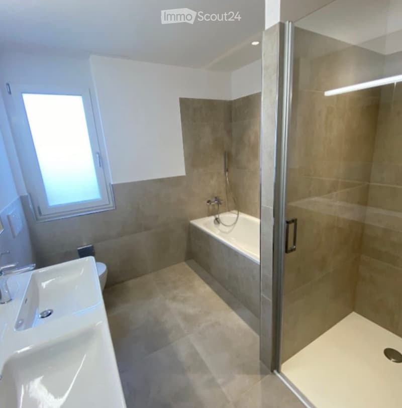 Modern bathroom with a bathtub, sink, toilet, and a glass-enclosed shower stall. The space features beige tiles and a large window providing natural light.