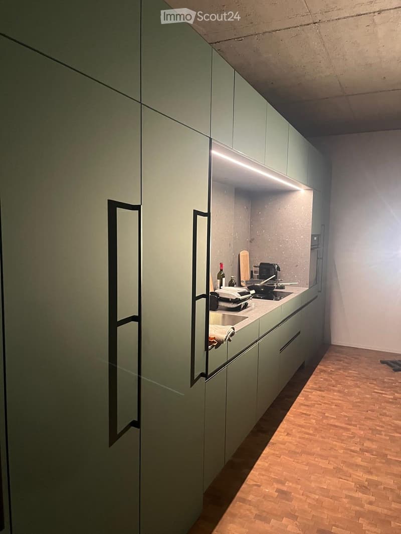 Modern kitchen with light green cabinets, wooden flooring, and built-in appliances including a sink, stove, and refrigerator. A bottle and cutting board are placed on the counter.