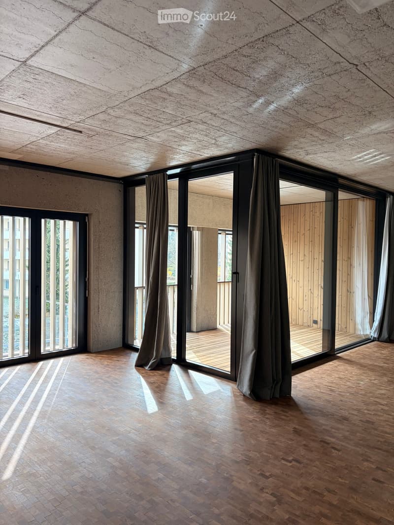 An unfurnished room with exposed concrete walls and ceiling, large glass sliding doors leading to a wooden decked balcony area, and wooden flooring.