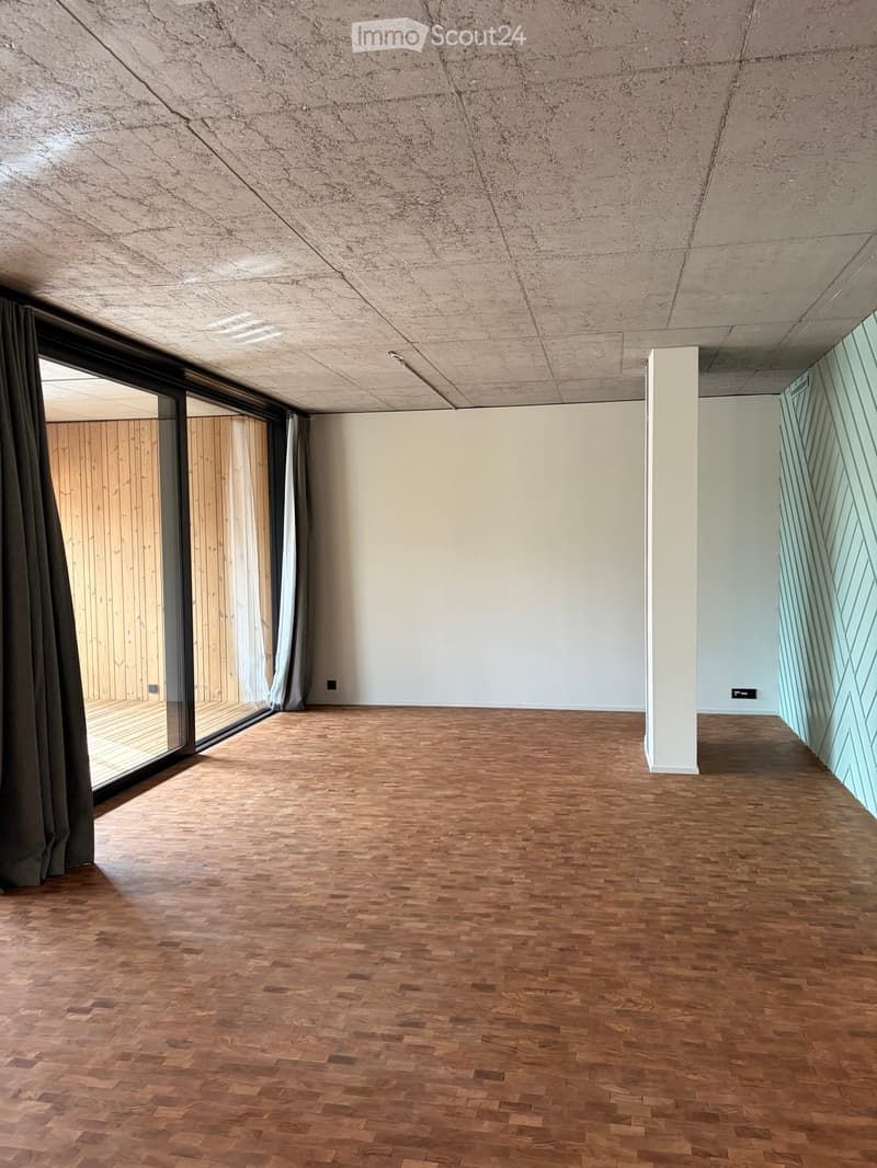 Empty room with wooden flooring, white walls, and large glass windows with curtains. Exposed concrete ceiling and a white column visible.
