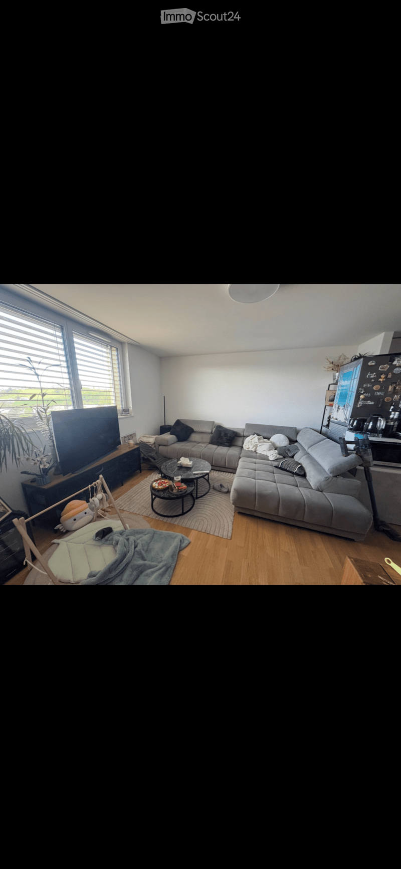 Modern living room with grey sectional sofa, coffee table, TV stand, and a small table with chairs. The room features wooden flooring, large windows with blinds, and a cozy rug.