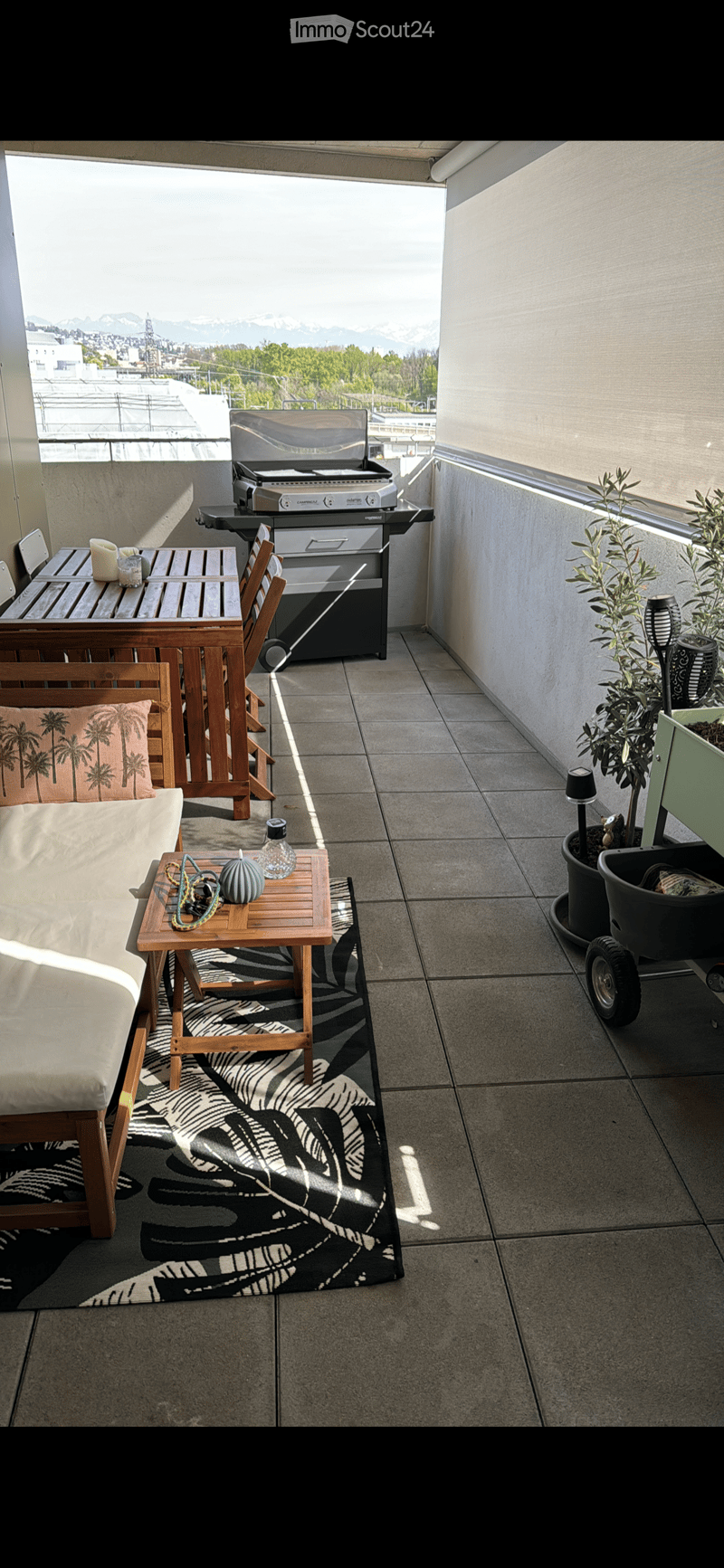 Balcony with a modern setup, featuring a grill, dining table, chairs, small sofa, and potted plants, overlooking a cityscape with mountains in the background.