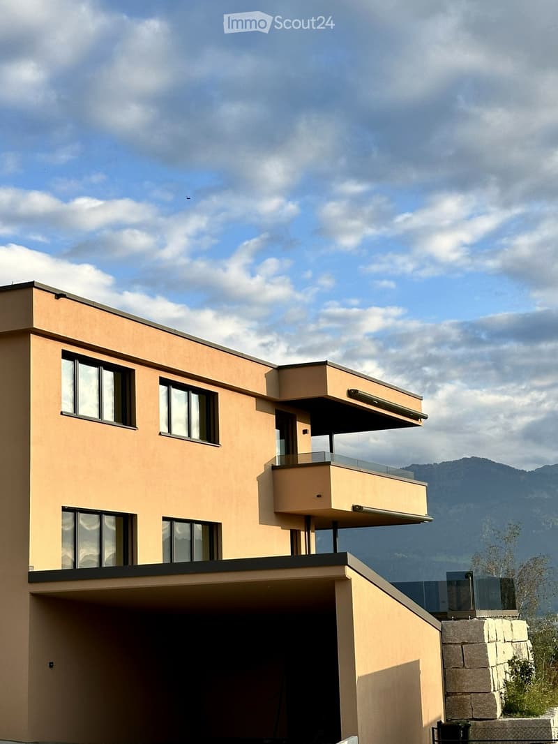 Modern two-story house with beige facade, large windows, and a balcony on the upper level. The house features a flat roof and is situated against a backdrop of mountains.
