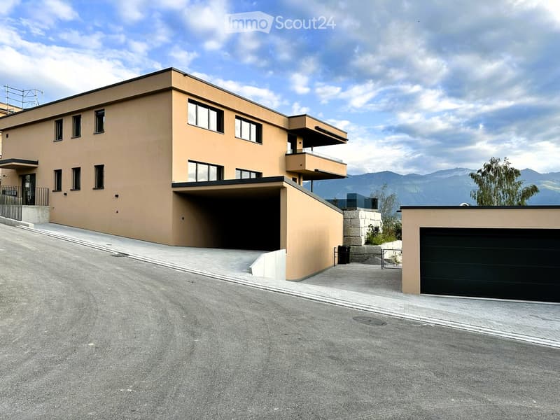Modern two-story house with beige facade, large windows, and a garage. The house features a raised ground floor and a balcony on the upper level.