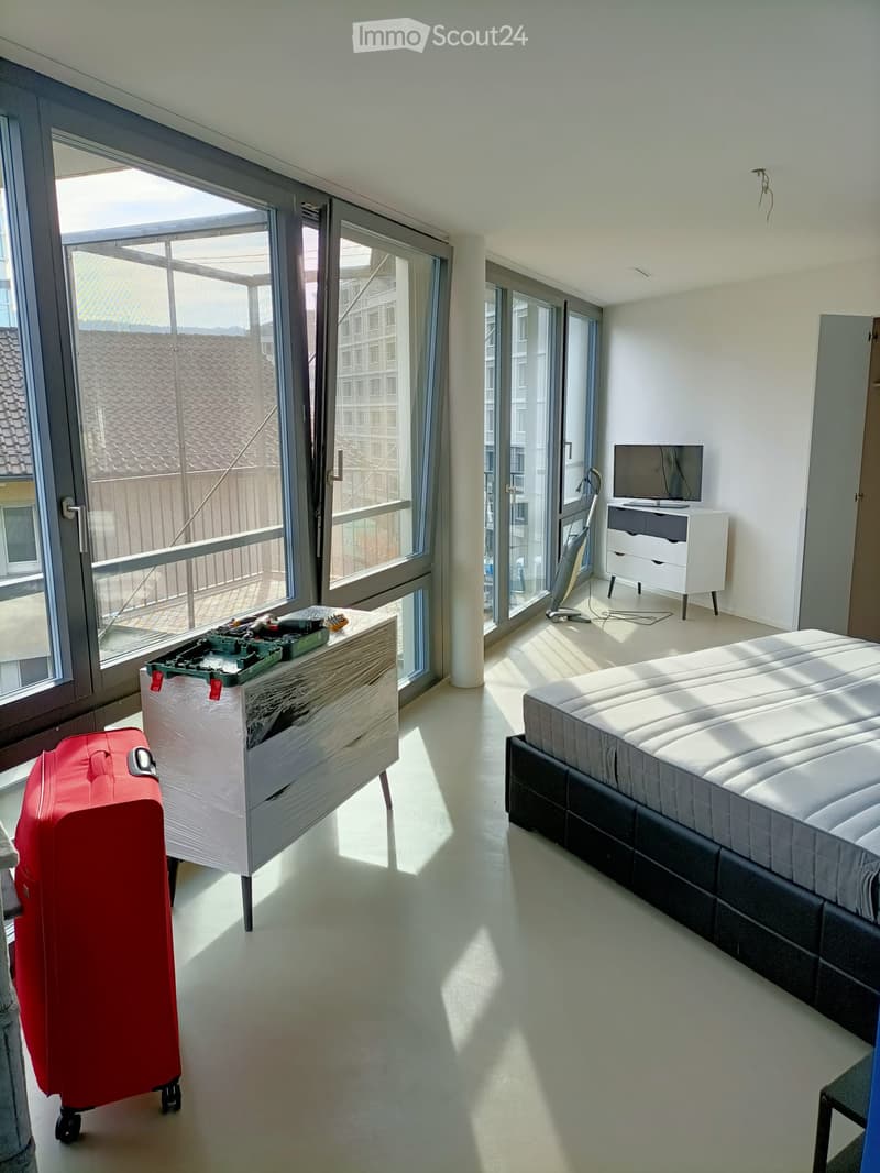 Bright bedroom with large windows and a balcony, featuring a bed, a table with tools, a red suitcase, and a TV stand.