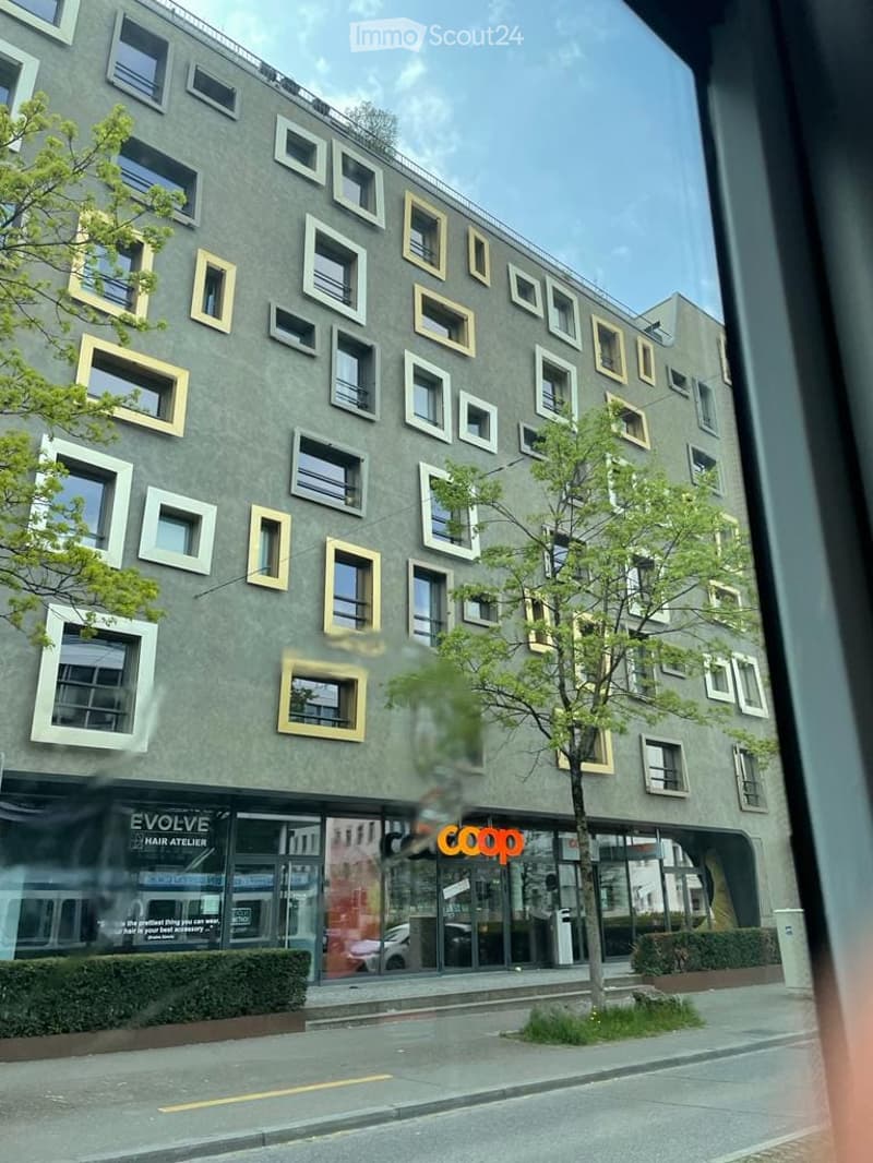 Modern multi-story building with grey facade and distinctive white-framed windows, featuring ground-floor storefronts including a hair salon and a Coop supermarket.