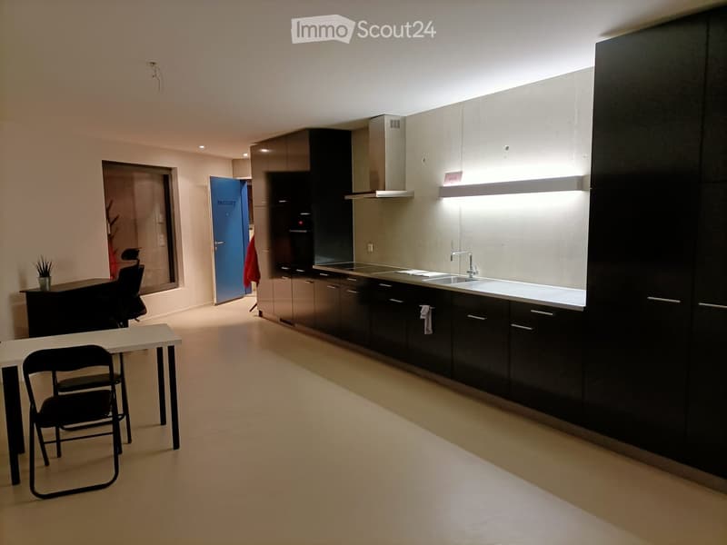 Modern kitchen with dark cabinets, stainless steel appliances, and a white countertop. A small table and chairs are placed in the foreground, and a door leads to another room.