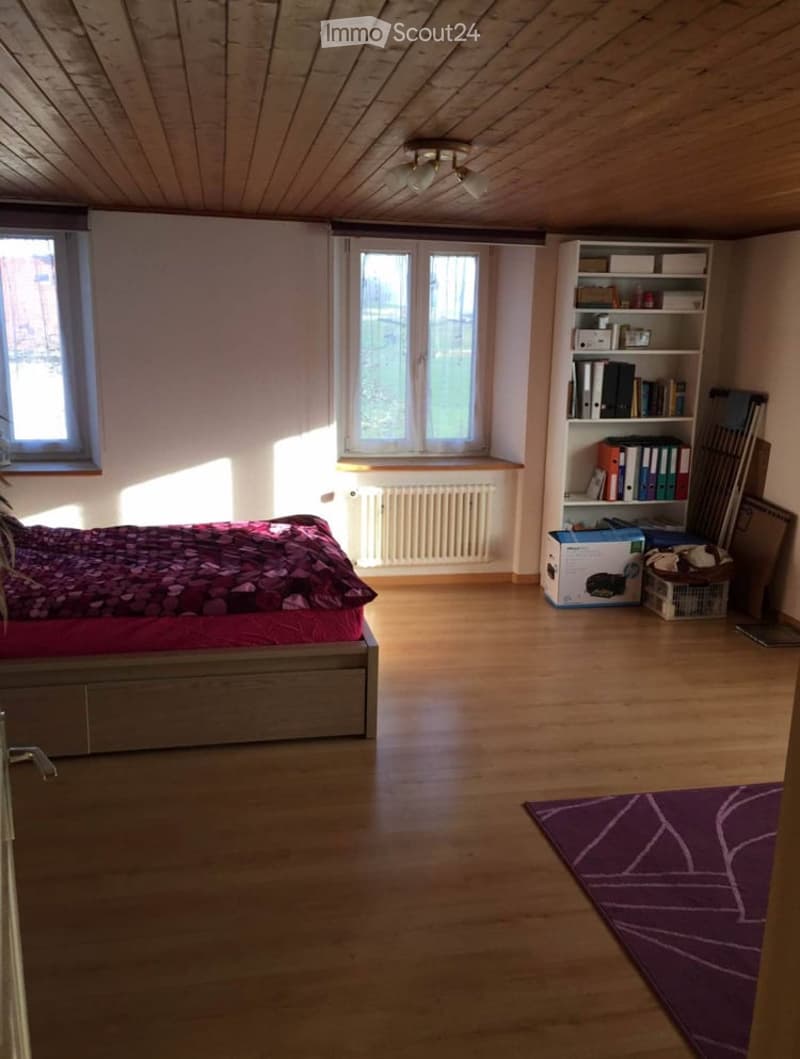 Bright bedroom with wooden flooring, large windows, a bed with red bedding, a shelf with books and storage boxes, and a purple rug.