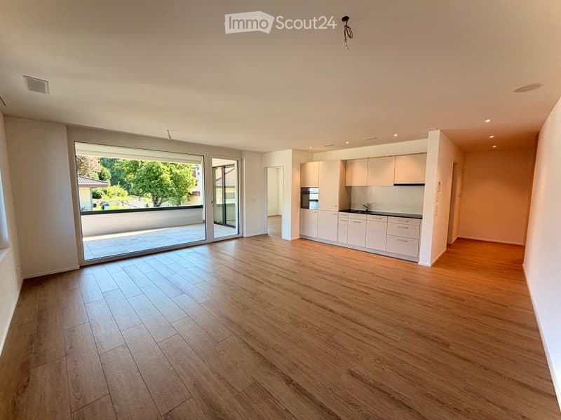 Open-plan unfurnished room with light wooden flooring, white kitchen cabinets, and large sliding glass doors leading to a balcony.