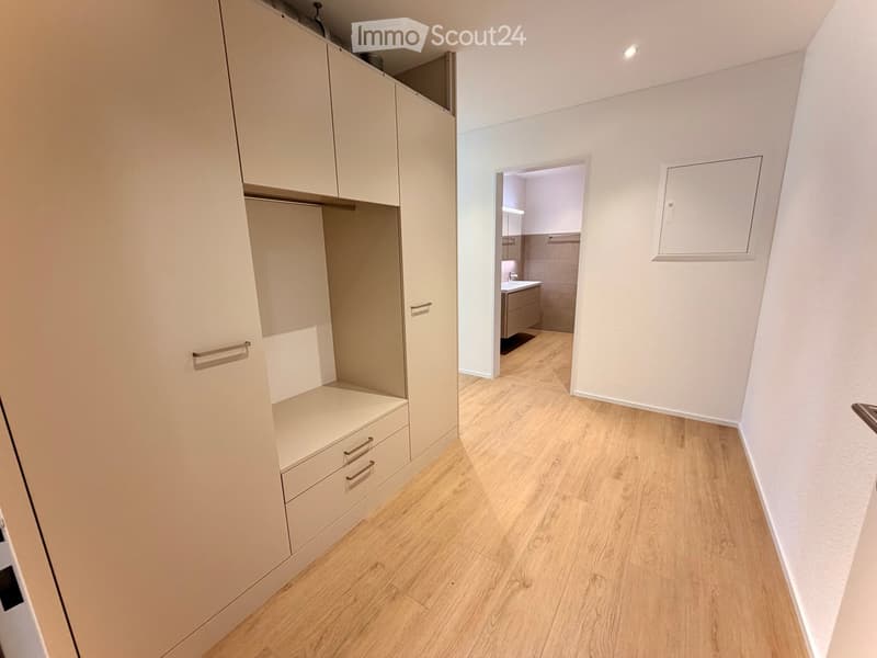 A clean, modern hallway with light wooden flooring and white walls. A built-in wardrobe with drawers and shelves is on the left, and an open doorway leads to a small bathroom with a sink and tiled walls.