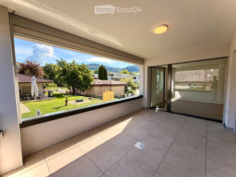 Spacious balcony with tiled floor, large glass sliding doors, and a scenic view of a green lawn, trees, and distant mountains.