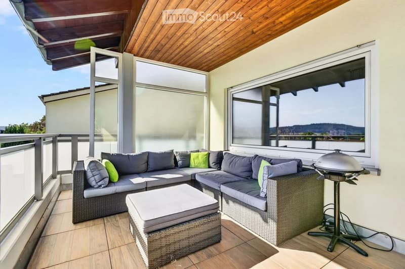 Balcony with a wooden ceiling, large windows, and a view of the landscape. Features a wicker sofa set with cushions, an ottoman, and a grill.