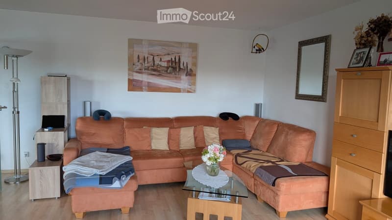 A modern living room with a large orange sectional sofa, a glass coffee table, a floor lamp, a mirror, and a wooden cabinet. The room features a painting on the wall and a vase with flowers on the coffee table.