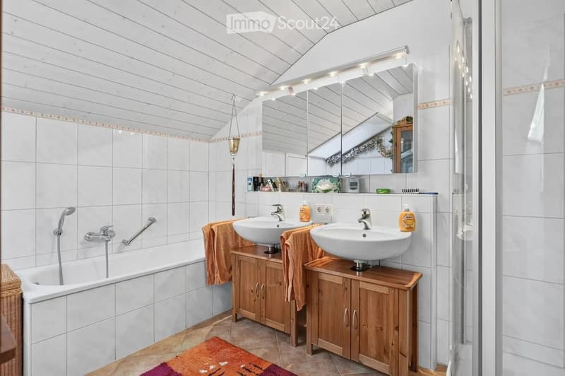Bathroom with white tiled walls, wooden cabinets, double sinks, a bathtub, and a shower area. Features include a large mirror with built-in lighting, towels, and a decorative rug on the floor.