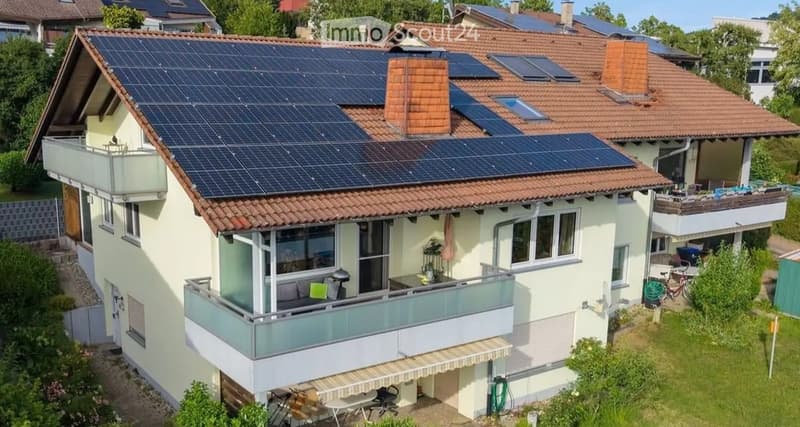 Two-story house with solar panels on the roof, multiple balconies, and surrounding greenery.