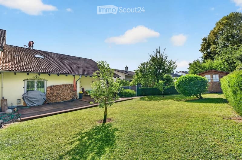Well-maintained garden with a wooden deck, covered grill, stack of firewood, small table and chairs, and a small shed.