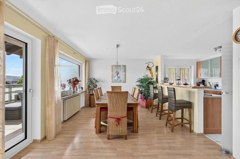 Bright, open-plan living and dining area with wooden flooring, large windows, a dining table with chairs, a kitchen island with stools, and potted plants.