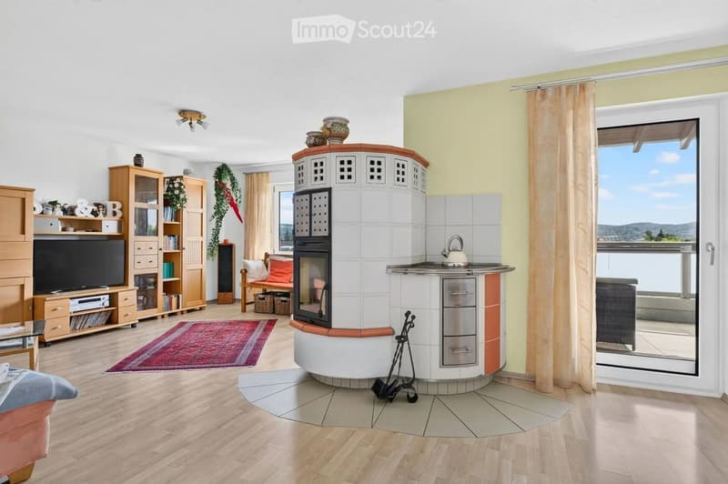 A modern living room with a central tiled fireplace, a sofa, a coffee table, a bookshelf, and a television. The room features large windows with curtains and a view of the outdoors.