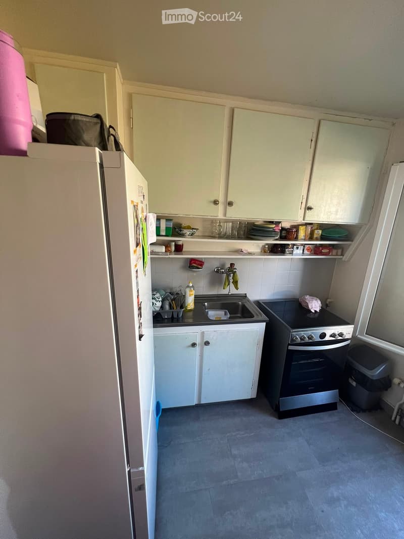 A compact kitchen with white cabinets, a stainless steel sink, a black oven, and a refrigerator. The countertops hold various kitchen items, and the floor is tiled.