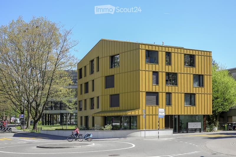 Modern yellow building with multiple windows and a flat roof, located on a street corner with trees and a cyclist passing by.