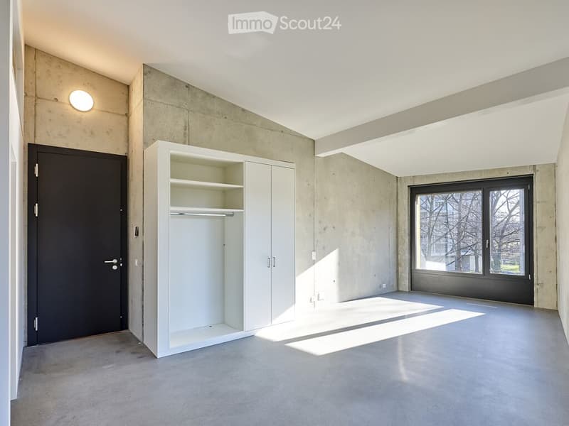 Empty room with concrete walls and floor, white built-in closet, large window, and a single light fixture on the wall.