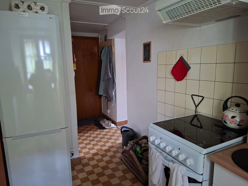 The kitchen features a white refrigerator, an induction stove with a floral teapot, and a white oven with control knobs. The floor is checkered, and there are towels hanging on the oven door and a broom in the corner.