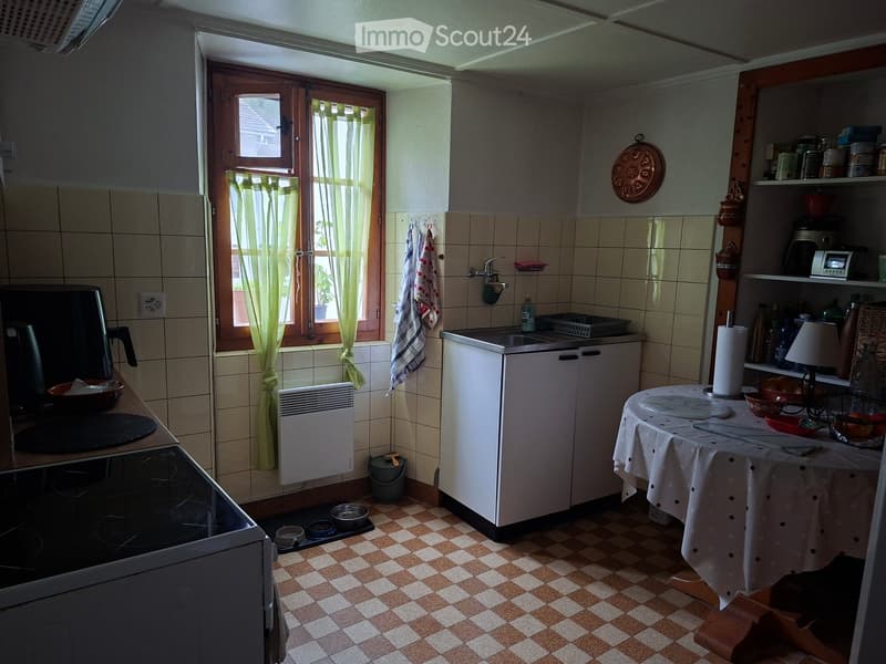 The kitchen features a checkered tile floor, white cabinets, a sink, a stove, and a small table with a tablecloth. There are various kitchen items on shelves and a window with green curtains.