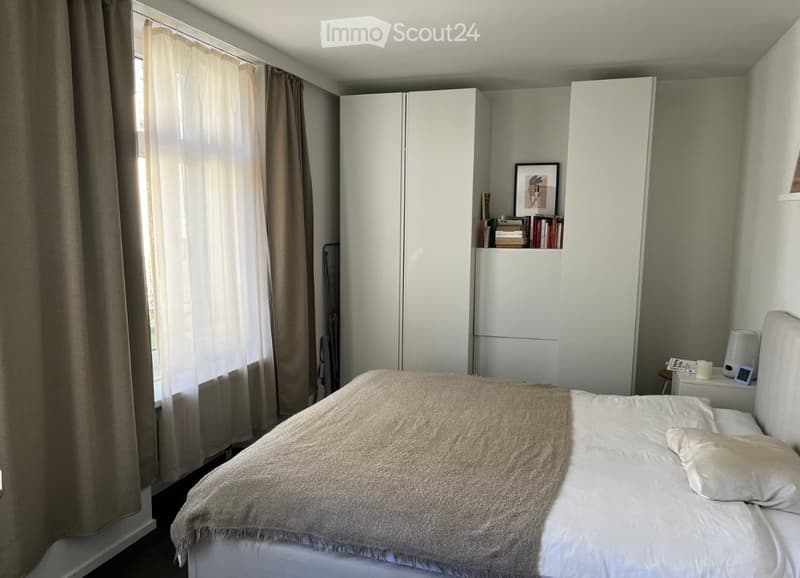 A modern bedroom with a large bed, nightstand, lamp, bookshelf, picture frame, and a tall white wardrobe. The room has large windows with light-colored curtains, allowing natural light to fill the space.