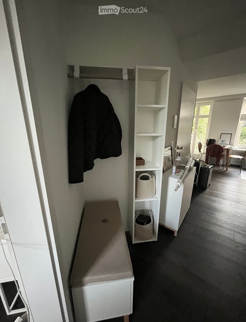 A small, furnished room with a bench, shelves, a chair, a table, and a suitcase visible.