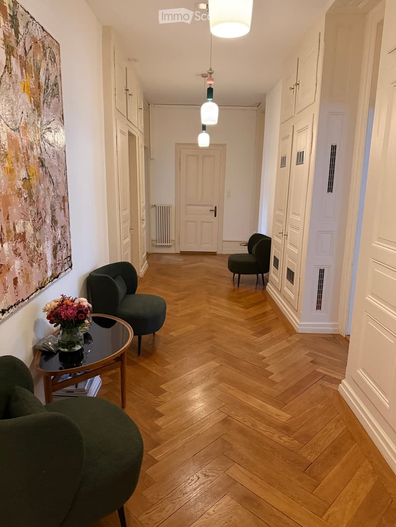 A hallway with wooden herringbone flooring, white walls, and several doors. There are green armchairs, a small round table with a vase of flowers, and a painting on the wall.