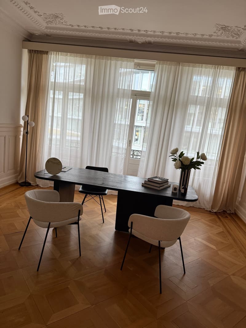 Elegant room with wooden floor, large windows with white curtains, a black table with chairs, a vase with white flowers, books, and a laptop on the table.